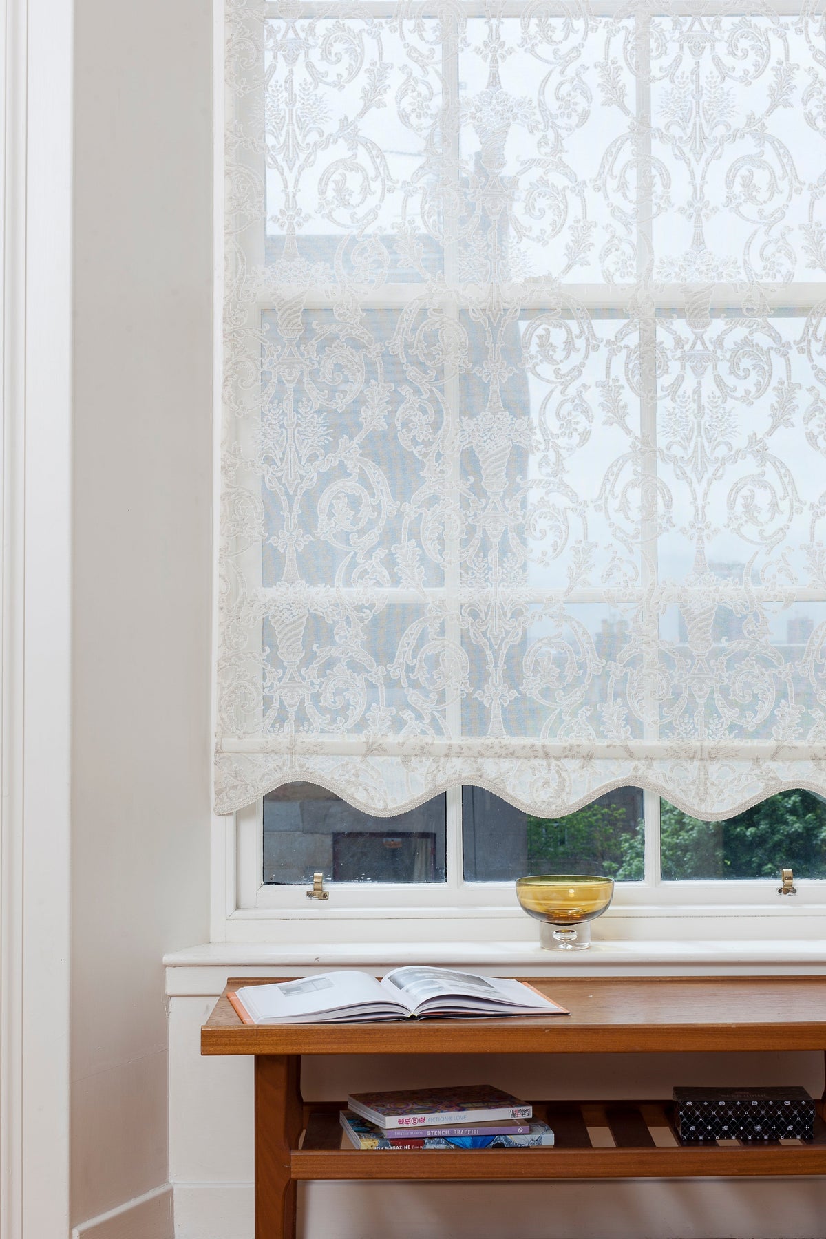 Custom roller shade in white Grecian scroll Madras lace with a scalloped hem installed on a window above a wooden table.