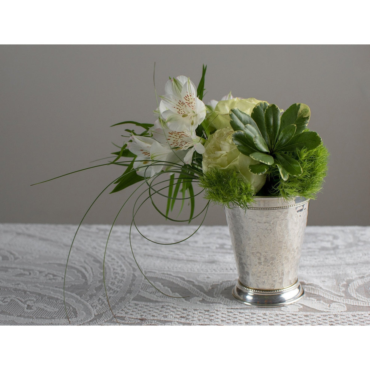 Hammered silver-plated mint julep cup with beaded rim holding a bouquet of white flowers on a lace tablecloth.