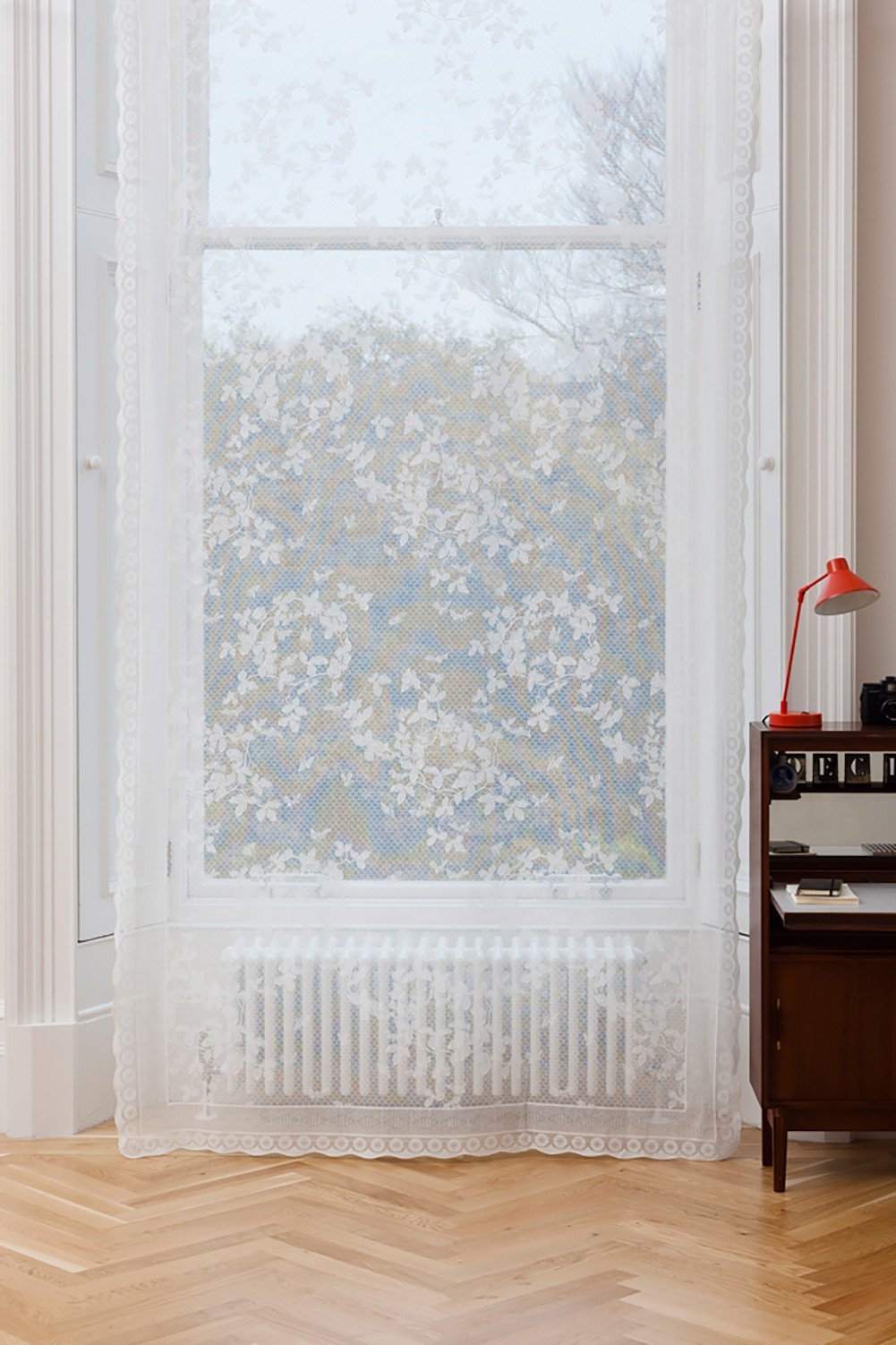 White cotton-blend lace curtain panel with intricate floral patterns, scalloped edges, and a rod pocket top on a window.