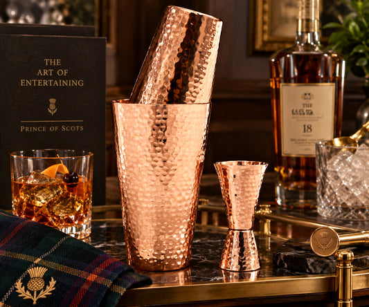 Hammered pure copper Boston cocktail shaker set with matching jigger on a bar cart with a glass of whiskey.