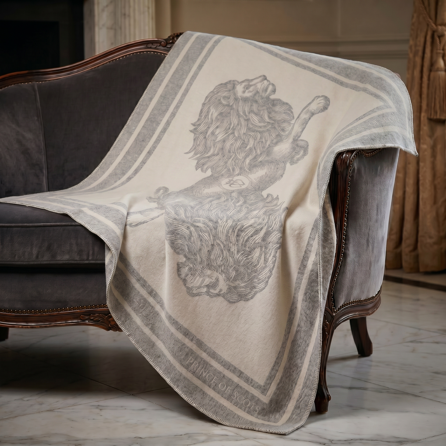 A cream-colored throw blanket with a gray lion illustration and striped border, draped over a dark gray velvet armchair in an elegant room.